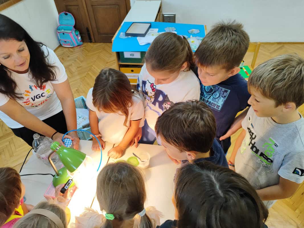 na fotografiji so učenci in učiteljica, ki stojijo za mizo in merijo temperaturo v posodi z namuzno svetilko.